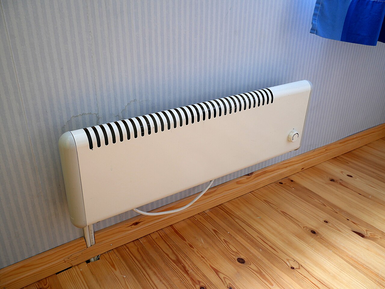 Radiador eléctrico blanco de pared con termostato, instalado junto al rodapié sobre suelo de madera